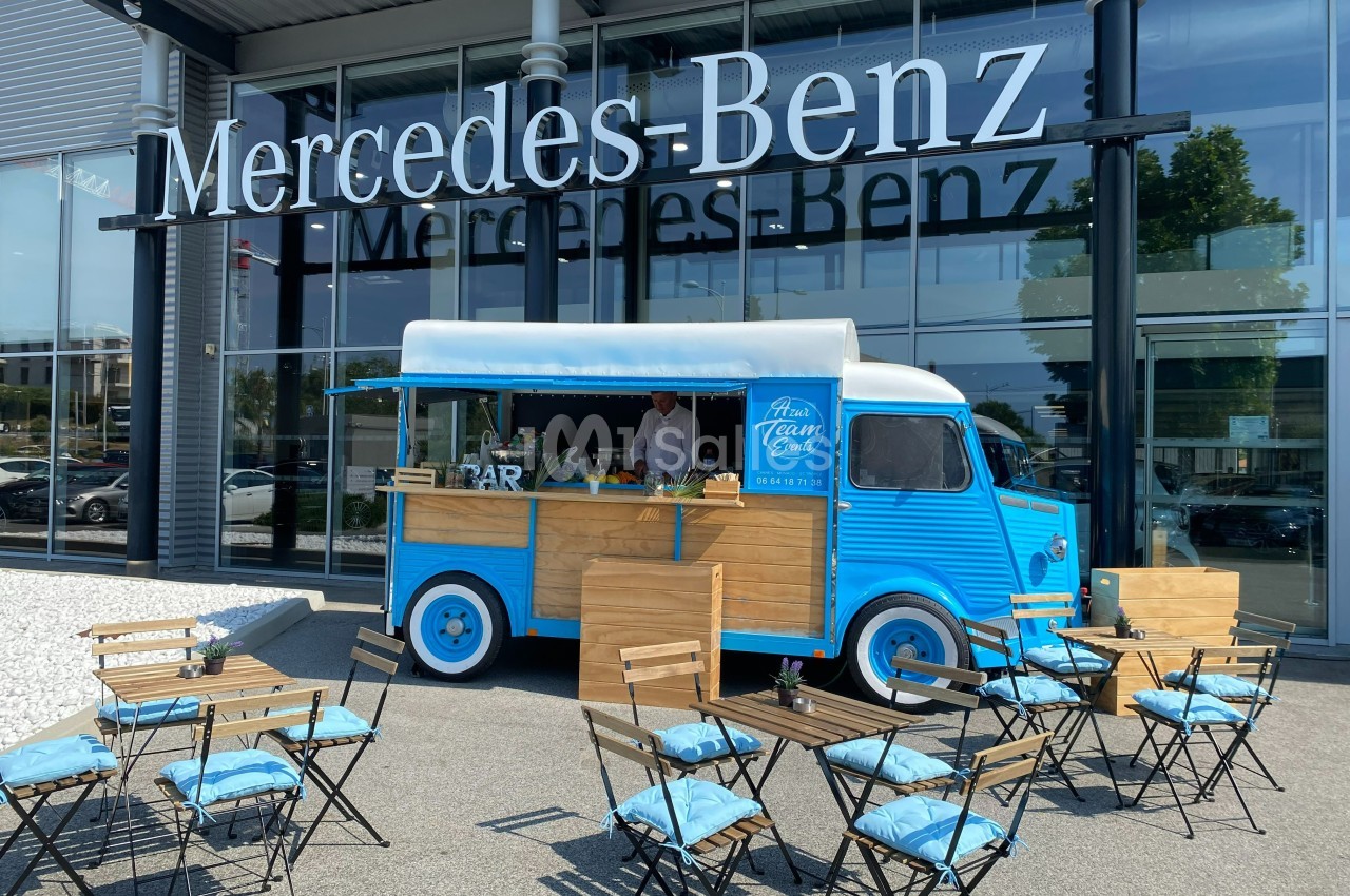 Camion-restaurant bleu vintage devant une concession Mercedes-Benz, avec tables et chaises disposées en terrasse.