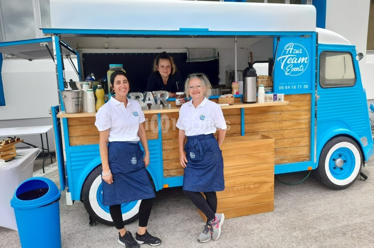 Un food truck bleu avec trois personnes souriantes en tablier devant un comptoir en bois.