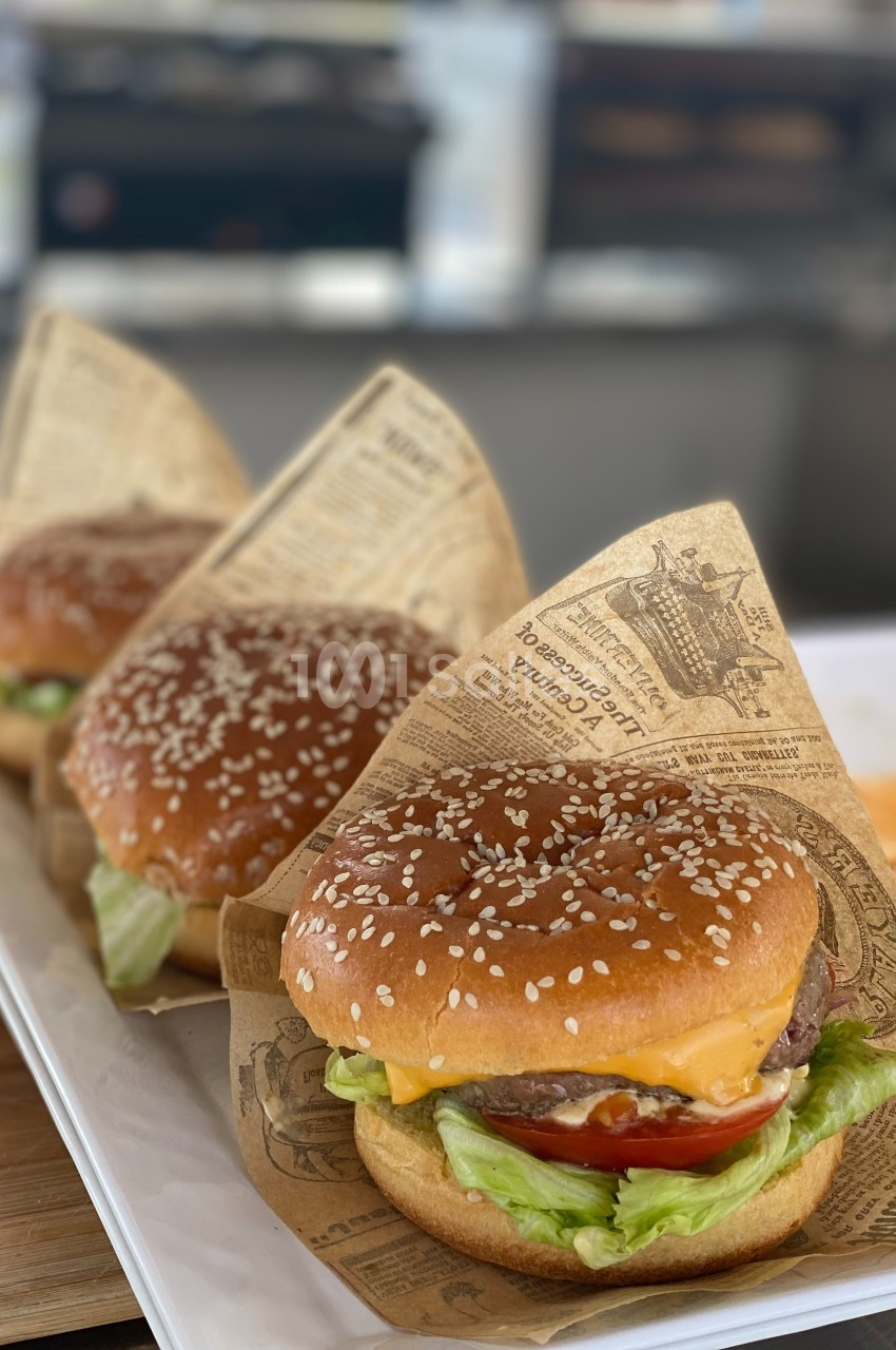 Trois hamburgers garnis de salade, tomate et fromage, enveloppés dans du papier imprimé, présentés sur un plateau blanc.