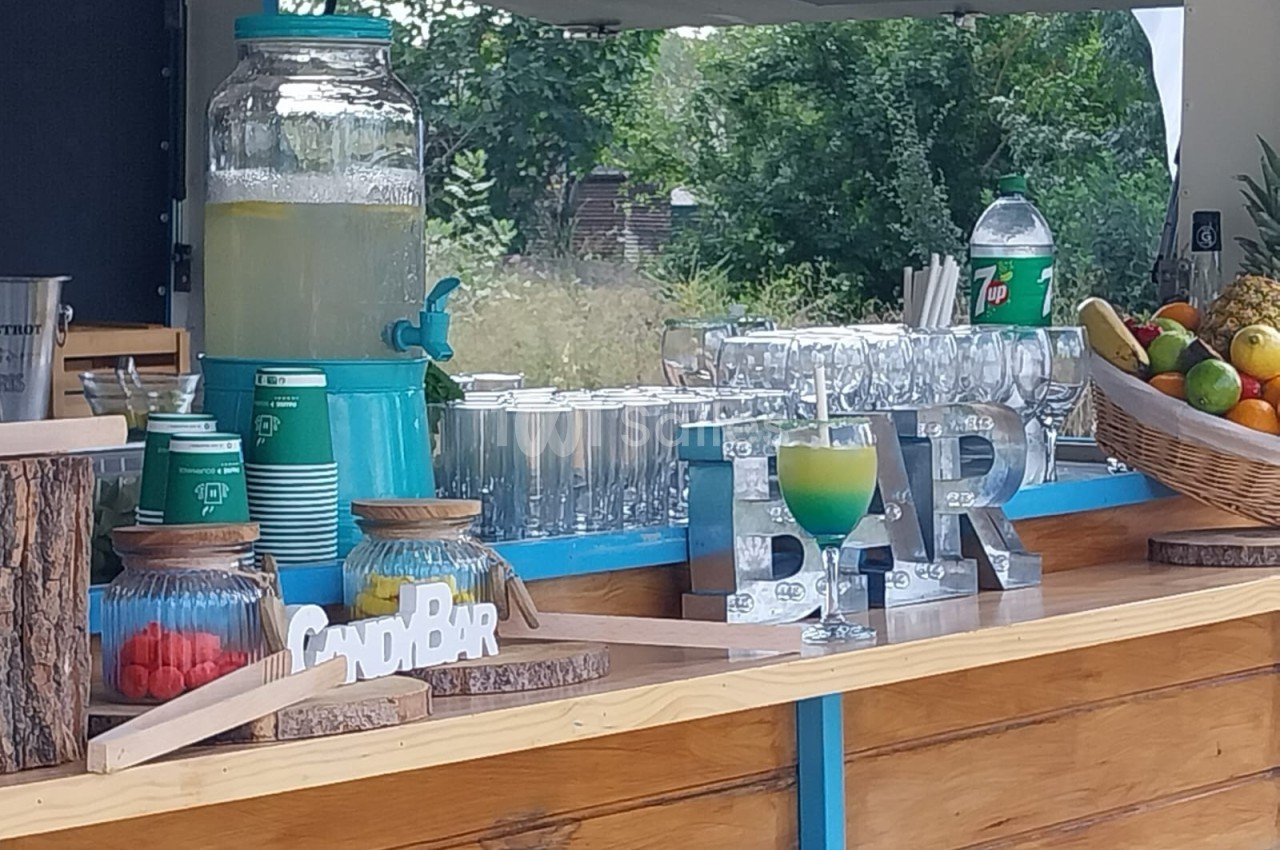 Stand en bois avec distributeur de limonade, bonbons, fruits frais et verres alignés sur un comptoir en extérieur.