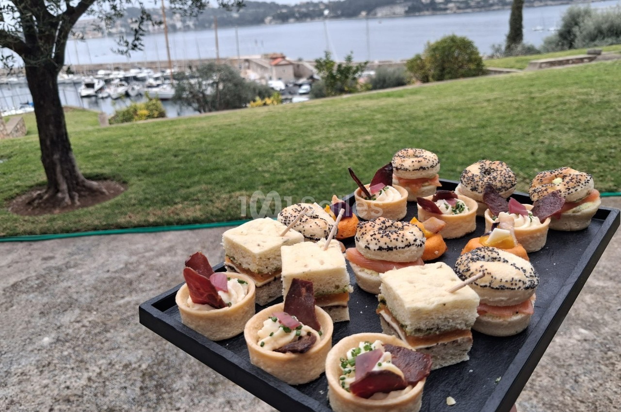Plateau de canapés variés présenté en extérieur avec vue sur un jardin, un port et une étendue d'eau.
