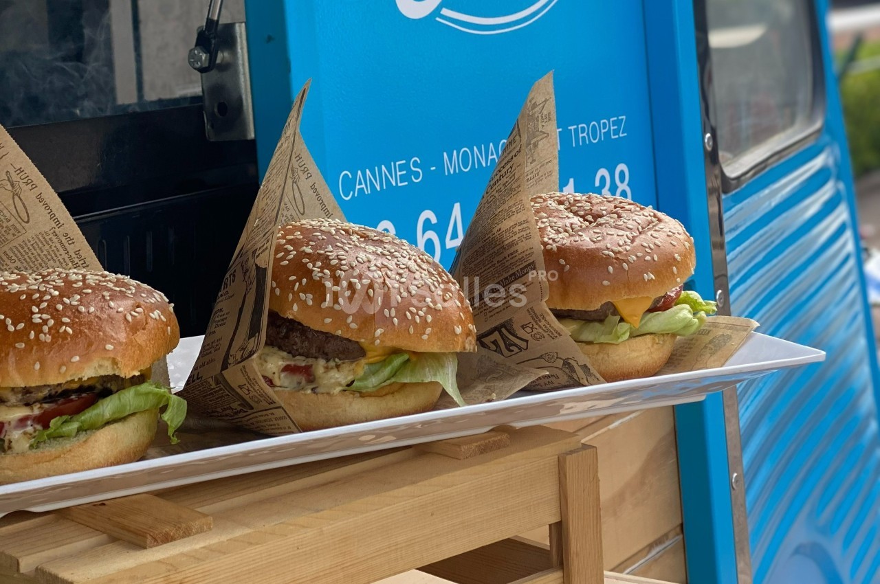 Trois hamburgers garnis de salade, fromage et steak, présentés sur un plateau devant un food truck bleu.