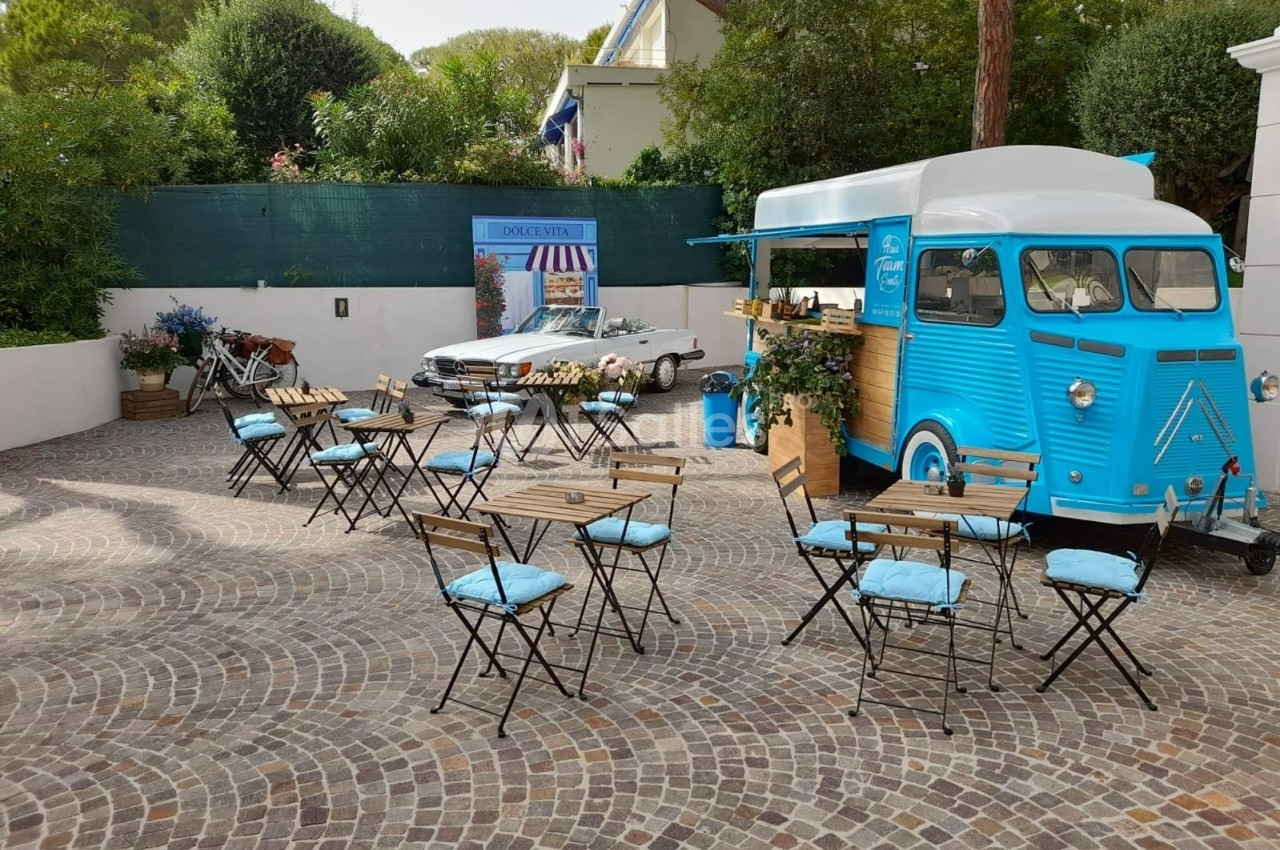 Camion bleu aménagé en café avec tables et chaises en terrasse sur une place pavée entourée de verdure.