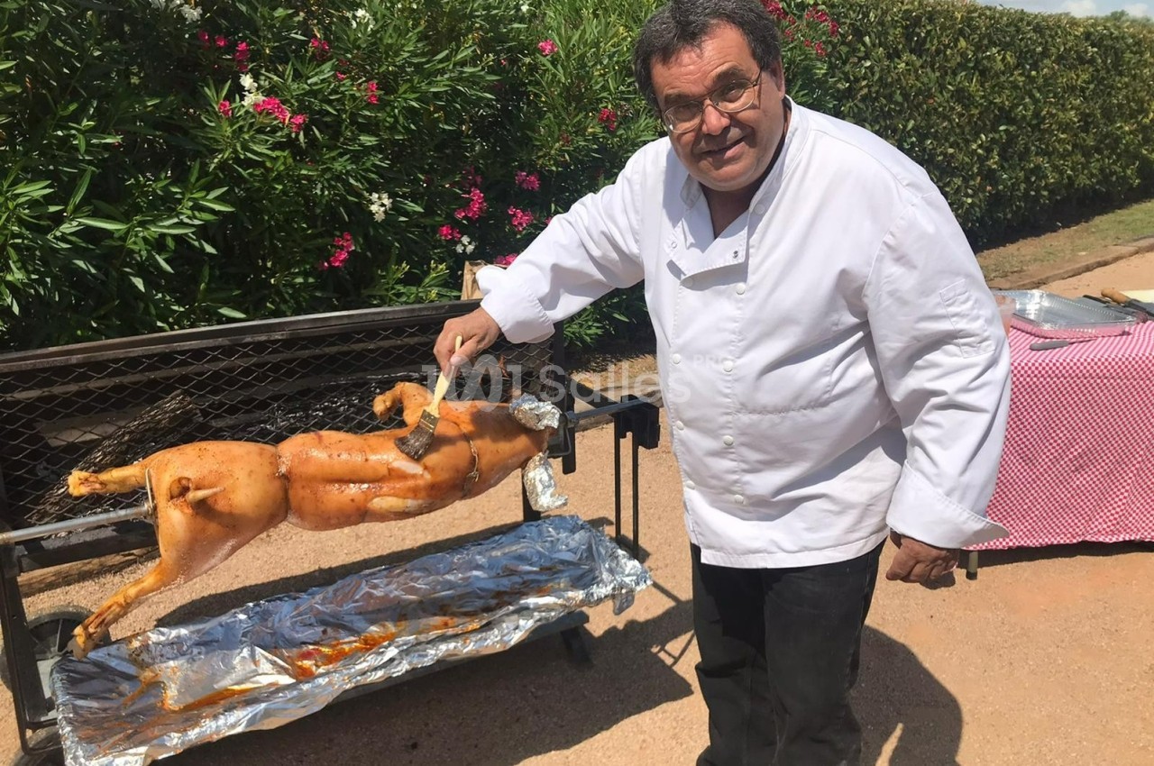 Un cuisinier badigeonne un cochon de lait rôti sur une broche devant un décor extérieur avec des arbustes fleuris.