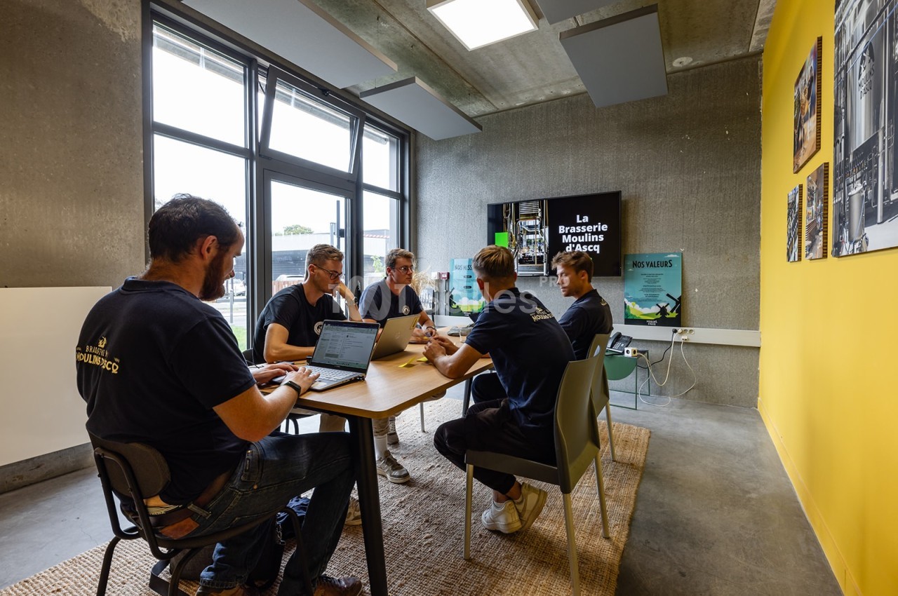 Un groupe de personnes discute autour d'une table dans une salle lumineuse avec des affiches et un écran en arrière-plan.