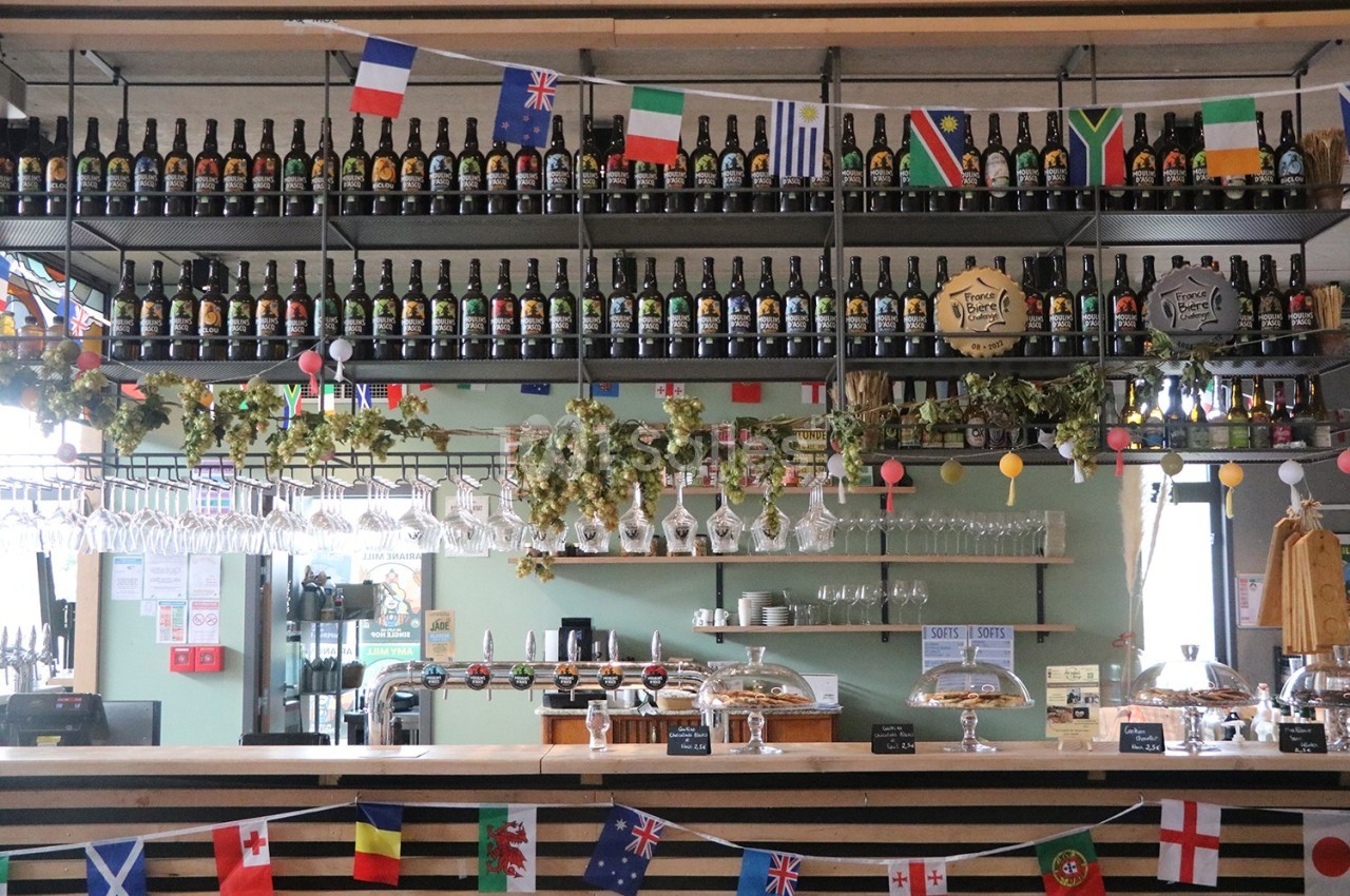 Bar décoré avec des drapeaux internationaux, des bouteilles alignées sur des étagères et des verres suspendus au plafond.