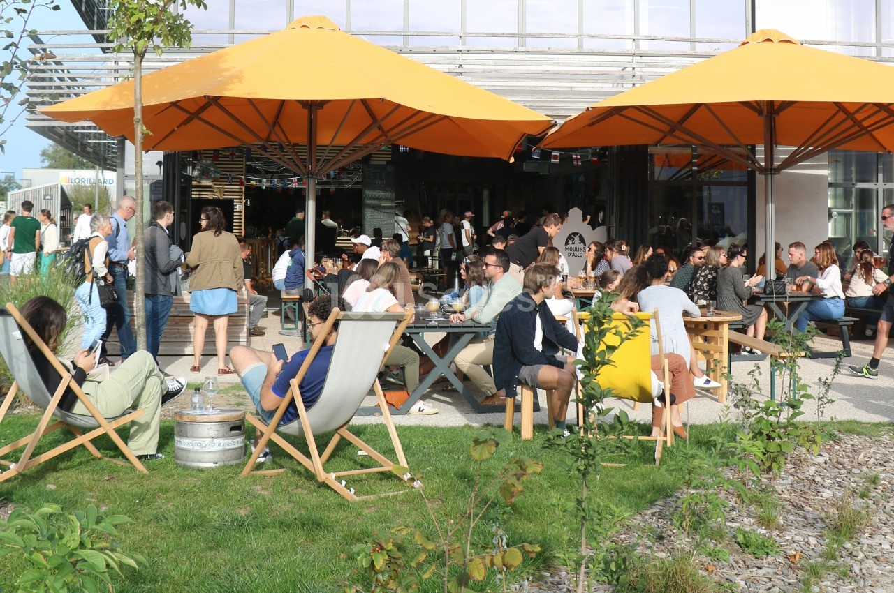 Personnes assises sous des parasols orange dans un espace extérieur, profitant d'un moment convivial par beau temps.