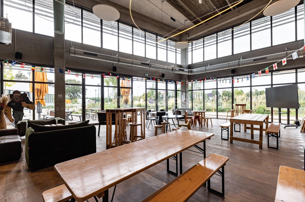 Salle lumineuse avec de grandes baies vitrées, tables en bois, chaises et décorations suspendues.