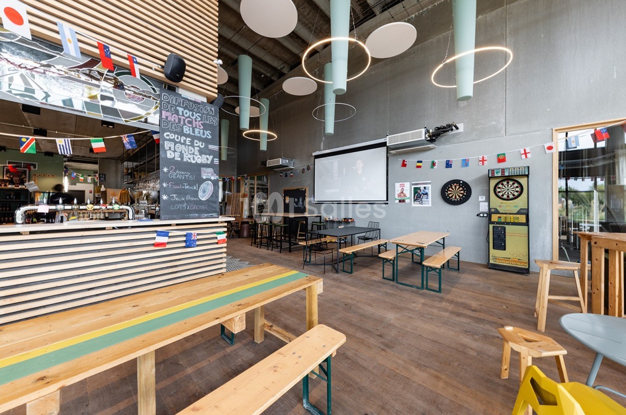Salle de bar avec tables en bois, écran de projection, cible de fléchettes et décorations aux couleurs internationales.