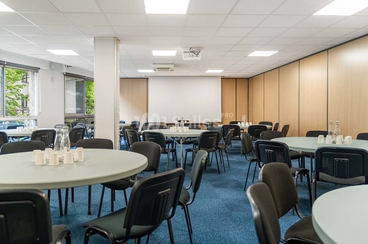 Salle de réunion avec tables rondes, chaises noires, lumière naturelle et projecteur au plafond.