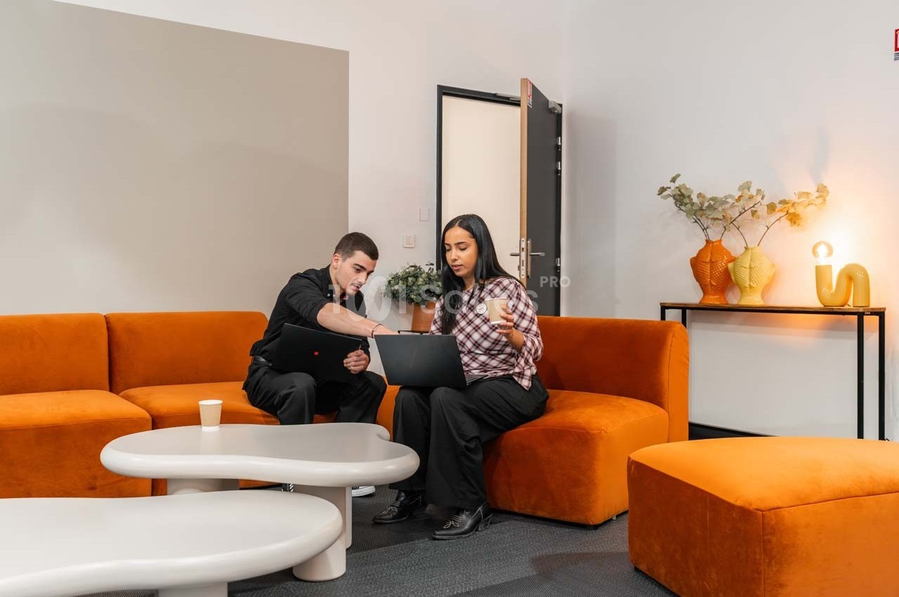 Deux personnes discutent en regardant un ordinateur portable dans un salon avec des canapés orange et une table basse.