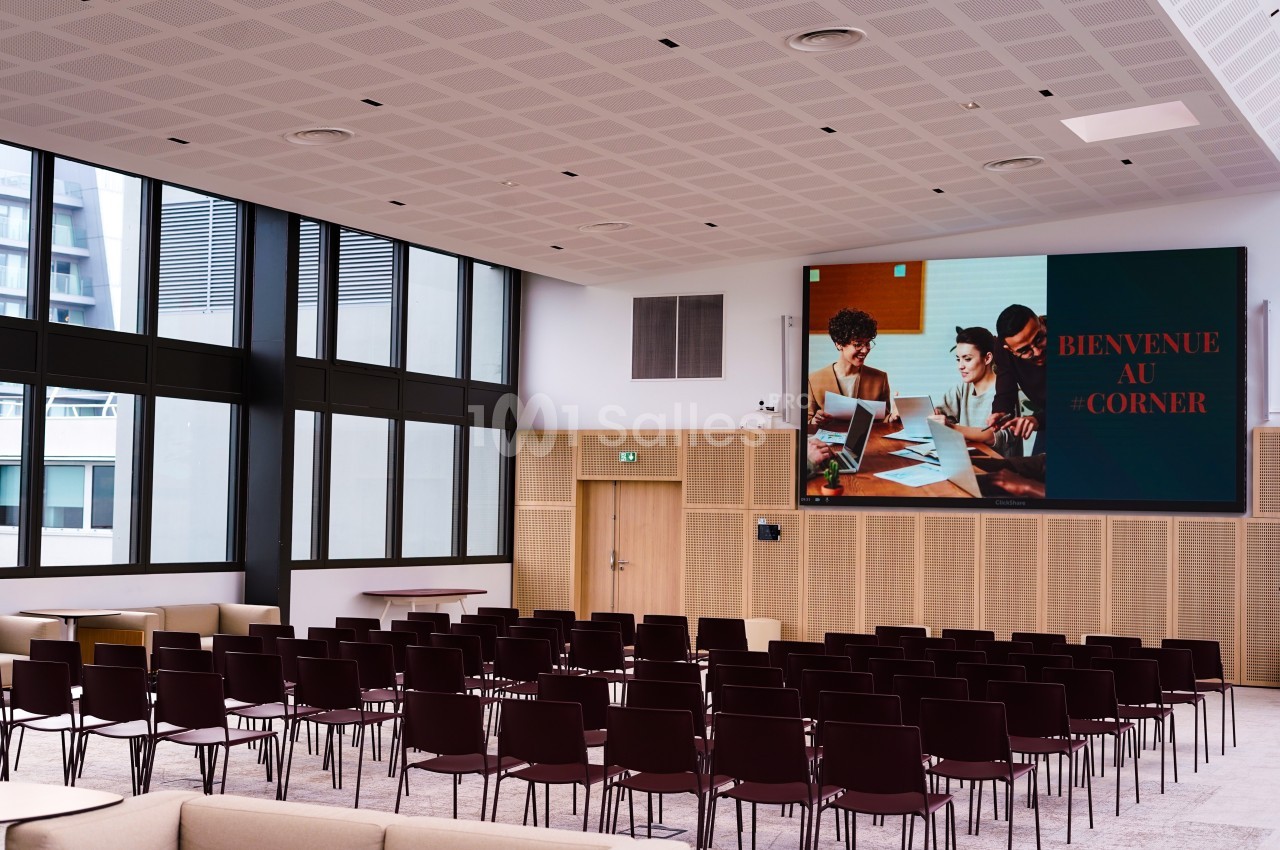 Salle de conférence moderne avec des rangées de chaises vides et un écran affichant ’Bienvenue au Corner’.