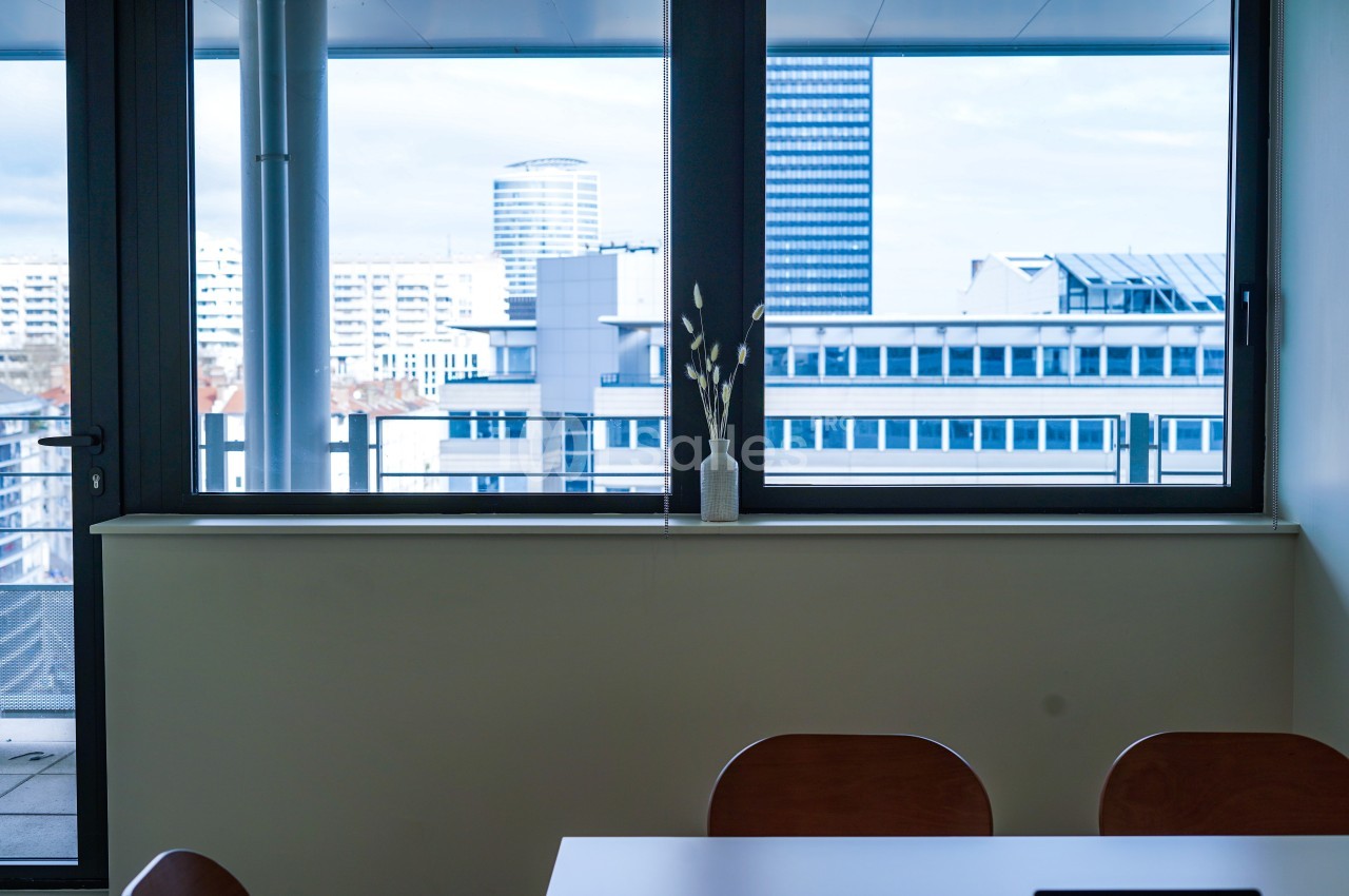 Vue depuis une salle de réunion avec une table, des chaises et une fenêtre donnant sur des immeubles modernes.