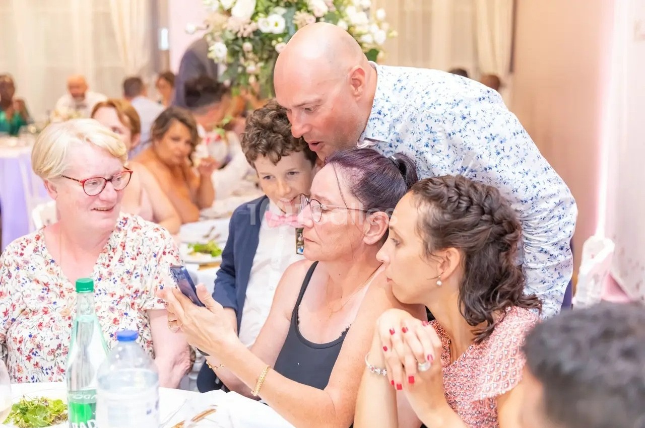 Un groupe de personnes assises à une table, regardant un téléphone, dans une ambiance festive avec des fleurs en arrière…