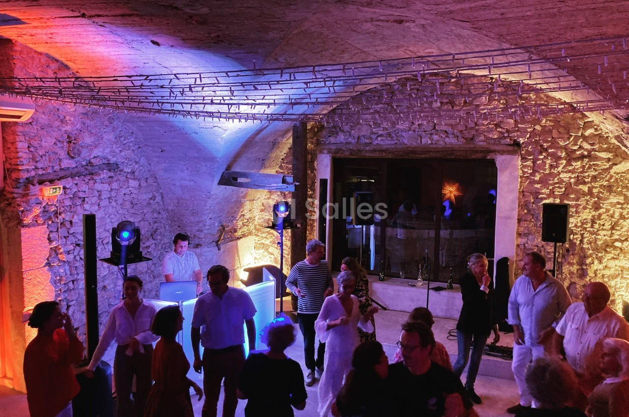 Personnes dansant et discutant dans une salle voûtée en pierre, éclairée par des lumières colorées, avec un DJ en arrière…