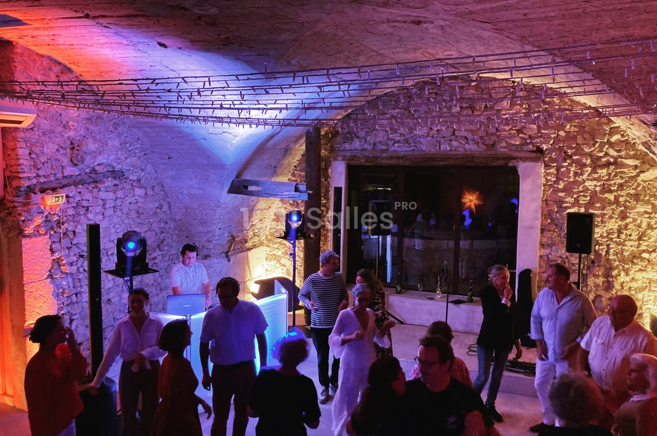 Personnes dansant dans une salle voûtée en pierre, éclairée par des lumières colorées, avec un DJ en arrière-plan.