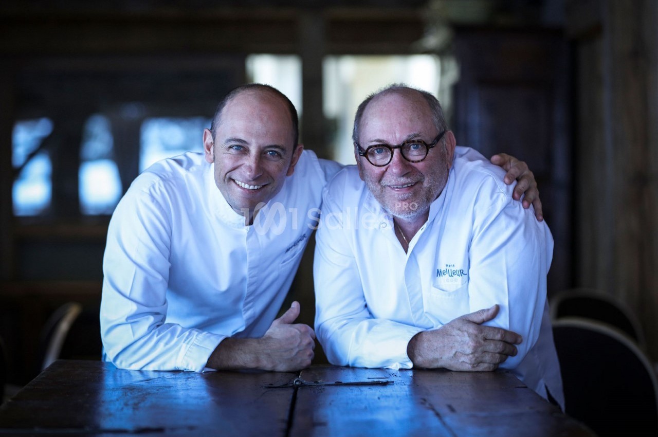 Deux chefs en tenue blanche sourient et posent côte à côte, assis à une table en bois dans un intérieur sombre.