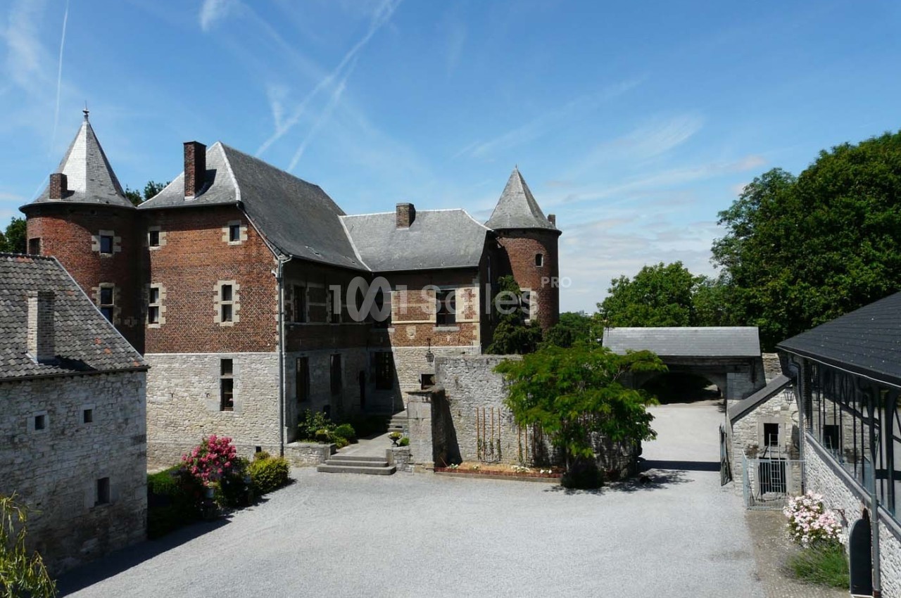 Château en pierre et briques rouges avec tours, cour pavée, arbres et ciel dégagé en arrière-plan.