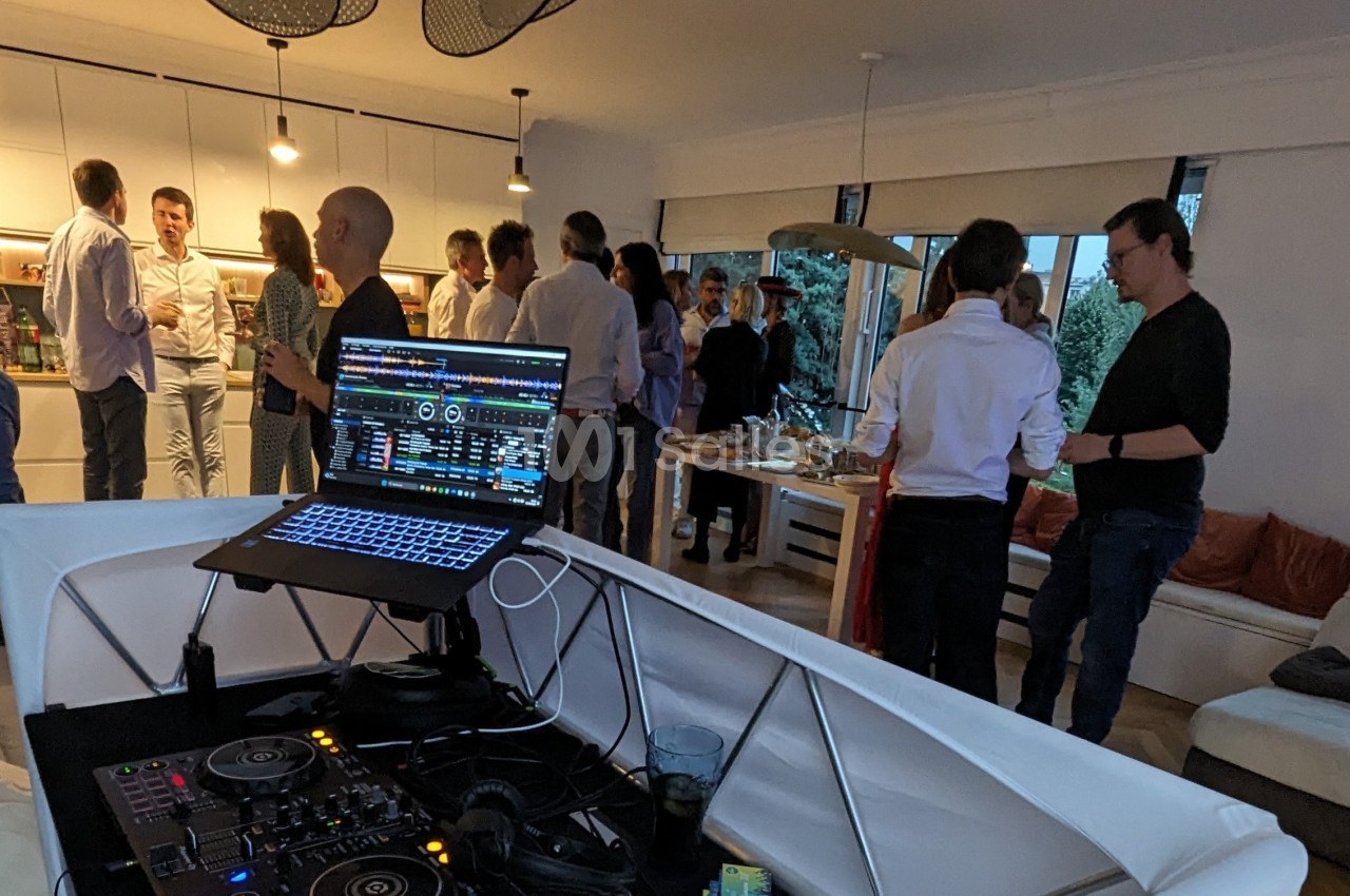 Des personnes discutent dans un salon lors d'une réception, avec une table de mixage et un ordinateur au premier plan.