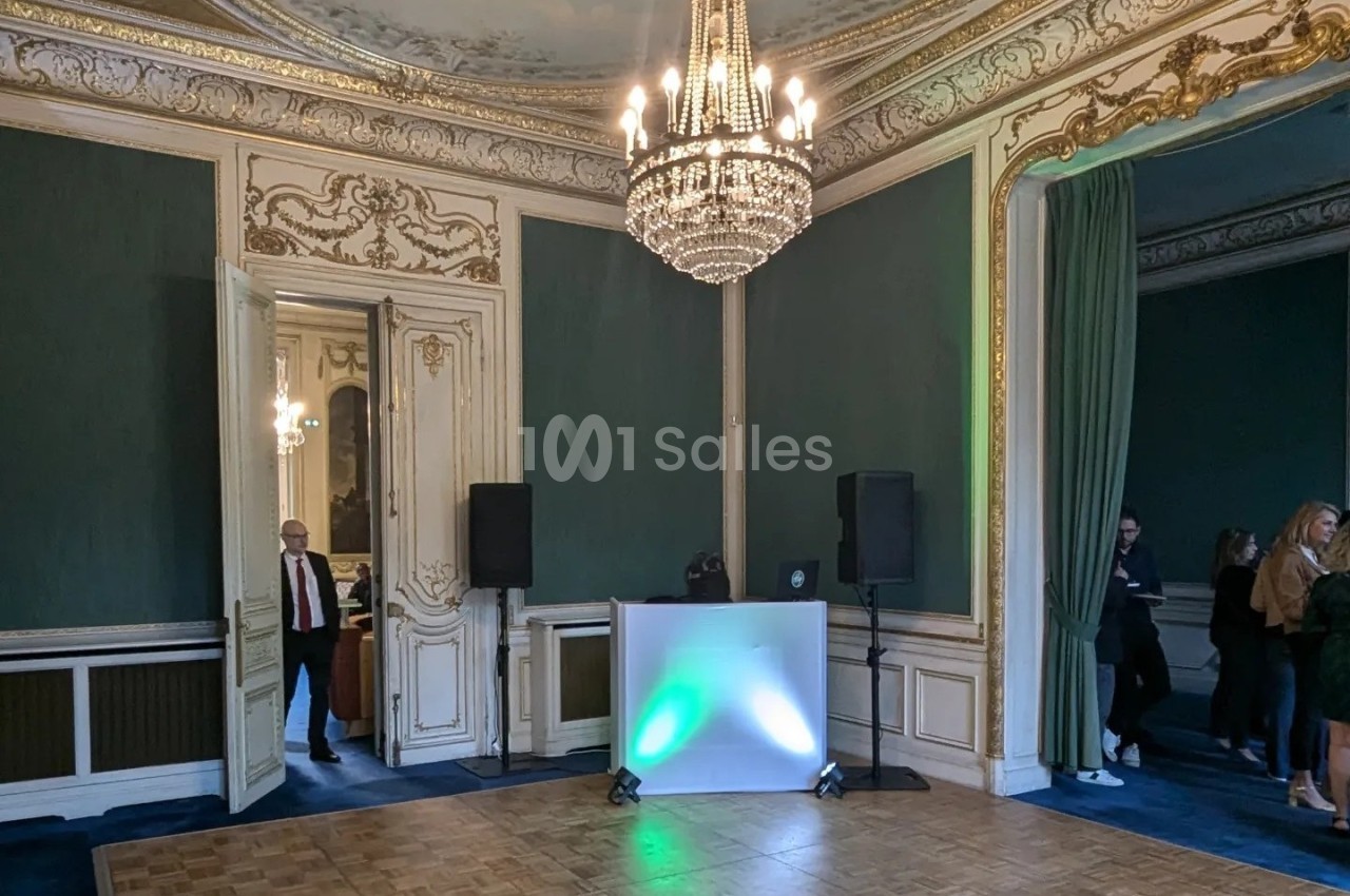 Salle élégante avec moulures dorées, murs verts, parquet, lustre central et équipement audio installé près d'une table…