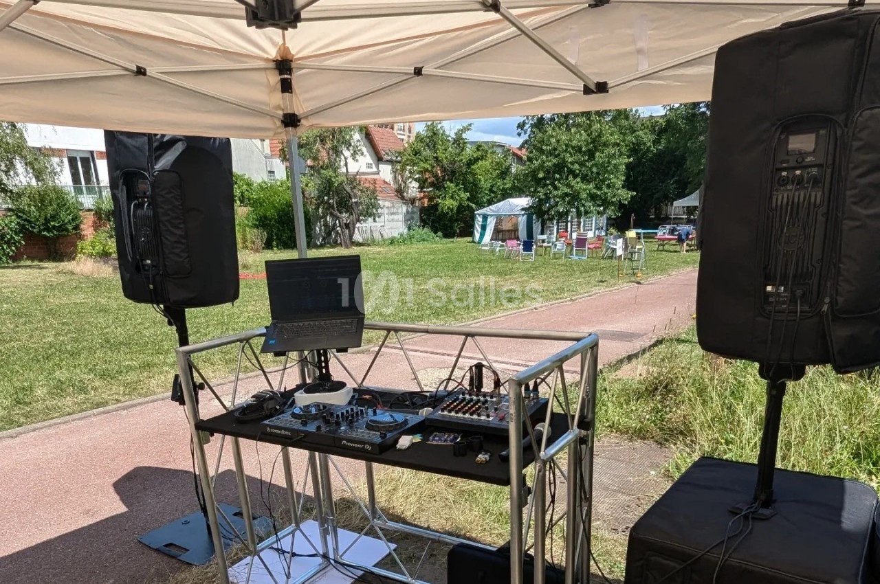 Équipement de sonorisation installé sous un abri, avec table de mixage, ordinateur et enceintes, dans un espace extérieur.
