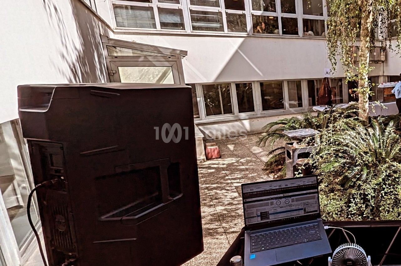 Poste de DJ en extérieur avec ordinateur portable, enceinte et végétation, devant un bâtiment moderne avec grandes fenêtres.