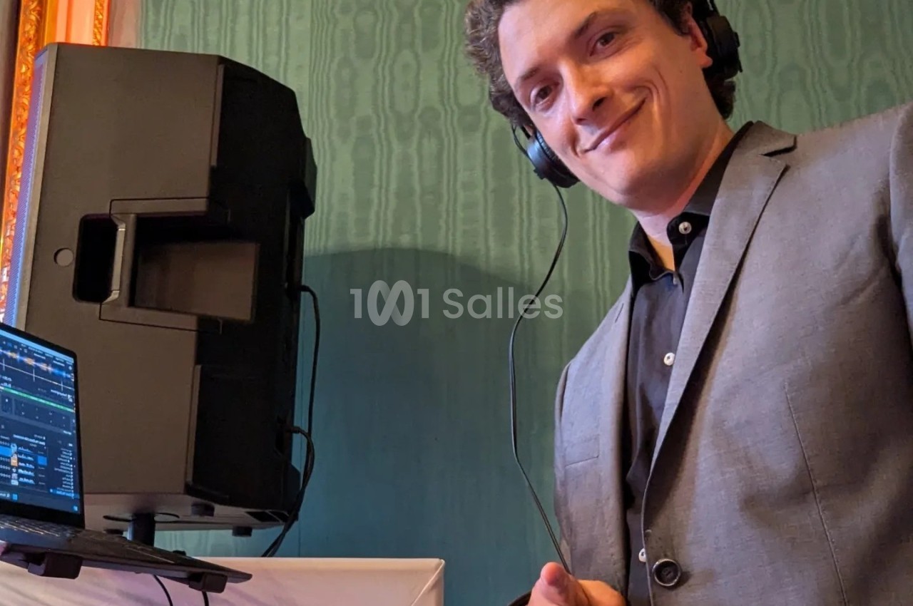 Un homme souriant portant un casque audio se tient près d'un équipement de DJ dans une pièce au mur vert.