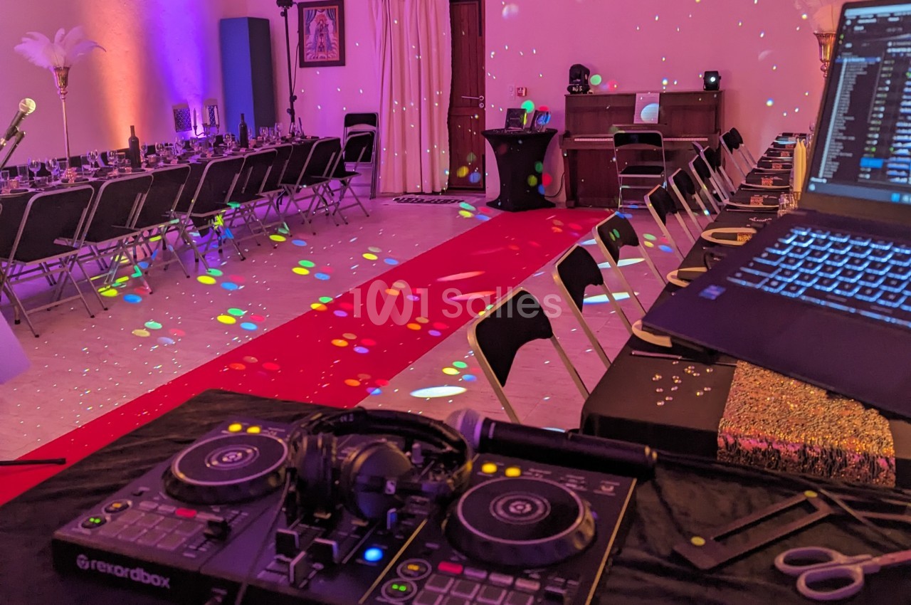 Salle décorée pour un événement avec tables dressées, éclairage coloré et matériel de DJ au premier plan.
