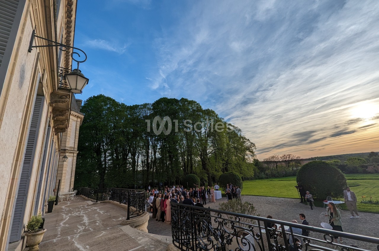Groupe de personnes rassemblées sur une terrasse devant un bâtiment historique, avec un parc et un ciel au coucher du soleil.