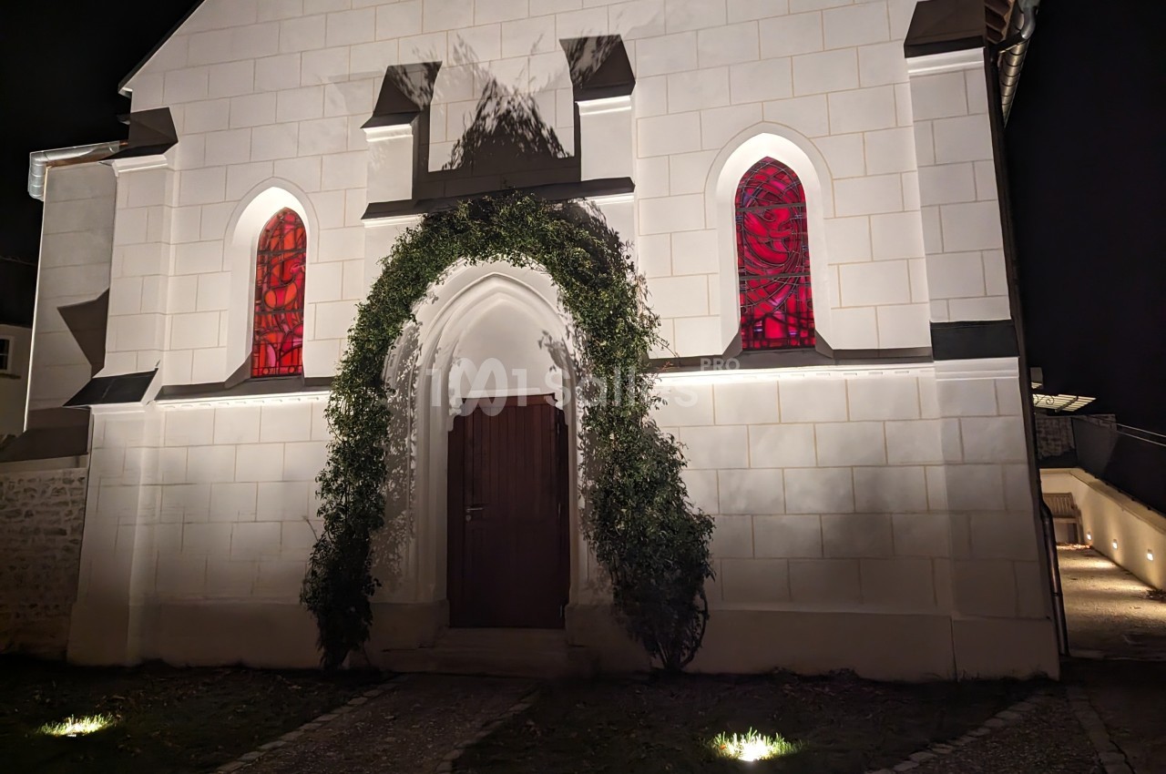Façade d'une petite chapelle blanche éclairée de nuit, avec une porte en bois entourée de verdure et des vitraux rouges.