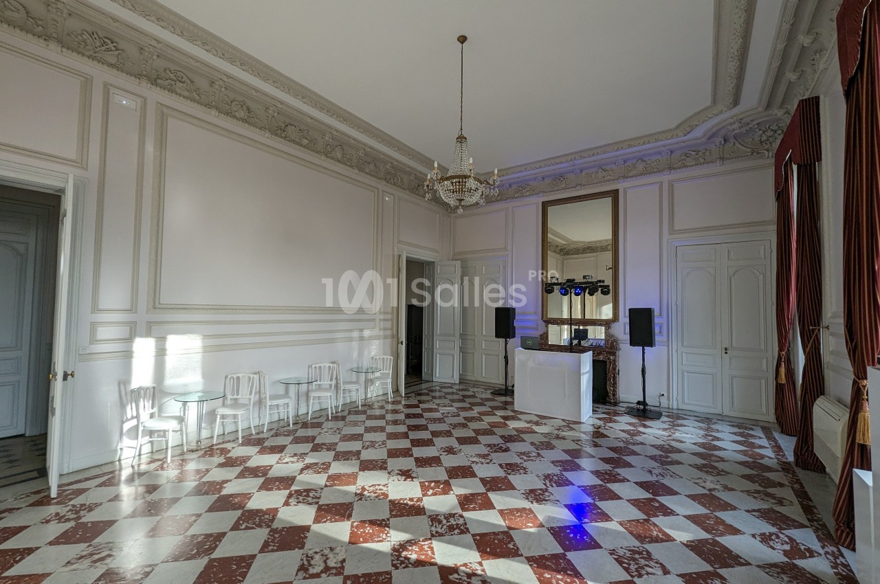 Salle lumineuse avec sol en damier rouge et blanc, moulures au plafond, lustre central et équipement de sonorisation.