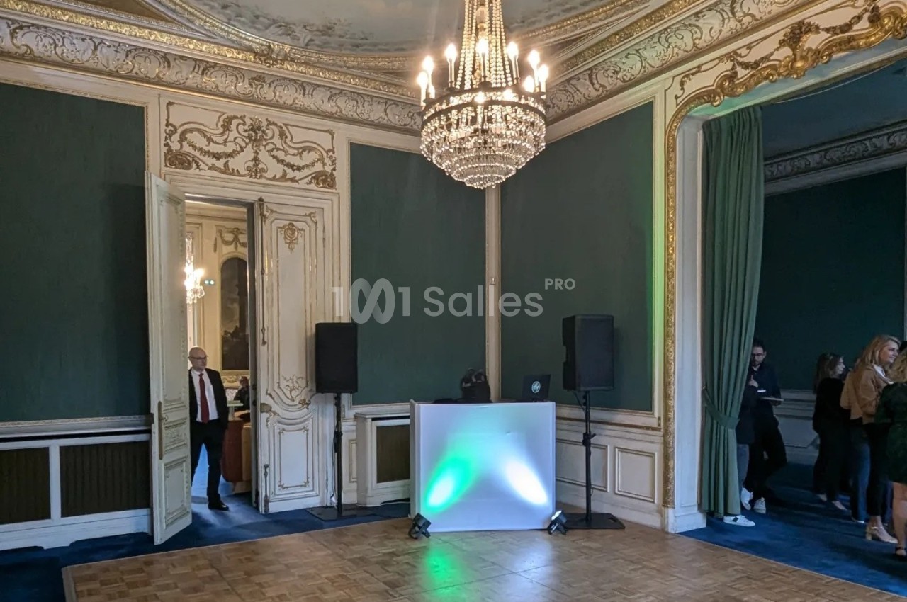 Salle élégante avec moulures dorées, lustre central, rideaux verts et un espace avec équipement de DJ.