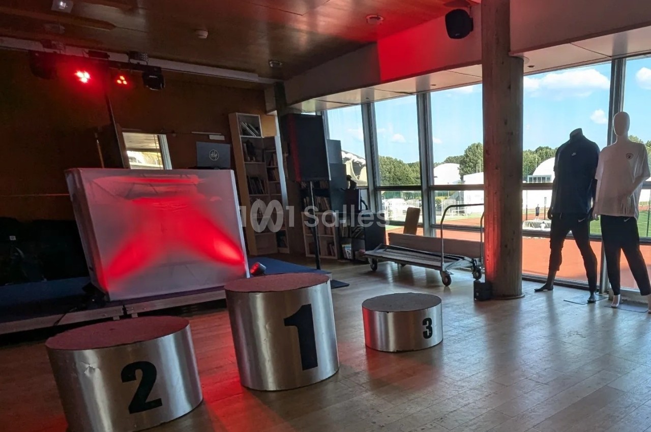 Podium métallique numéroté dans une salle lumineuse avec baies vitrées donnant sur une piste extérieure.