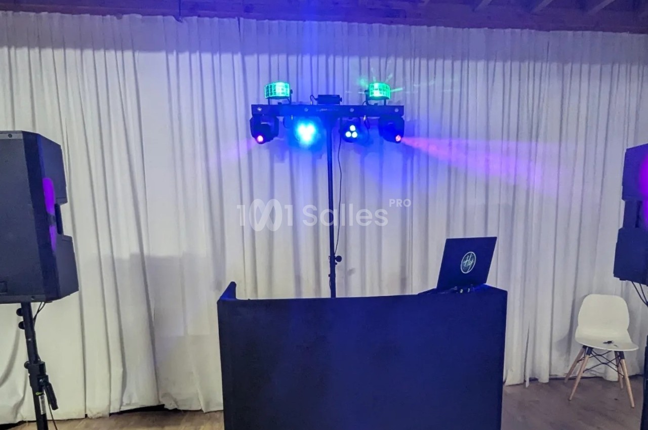 Poste de DJ avec table noire, ordinateur portable, enceintes et jeux de lumières colorées devant un rideau blanc.