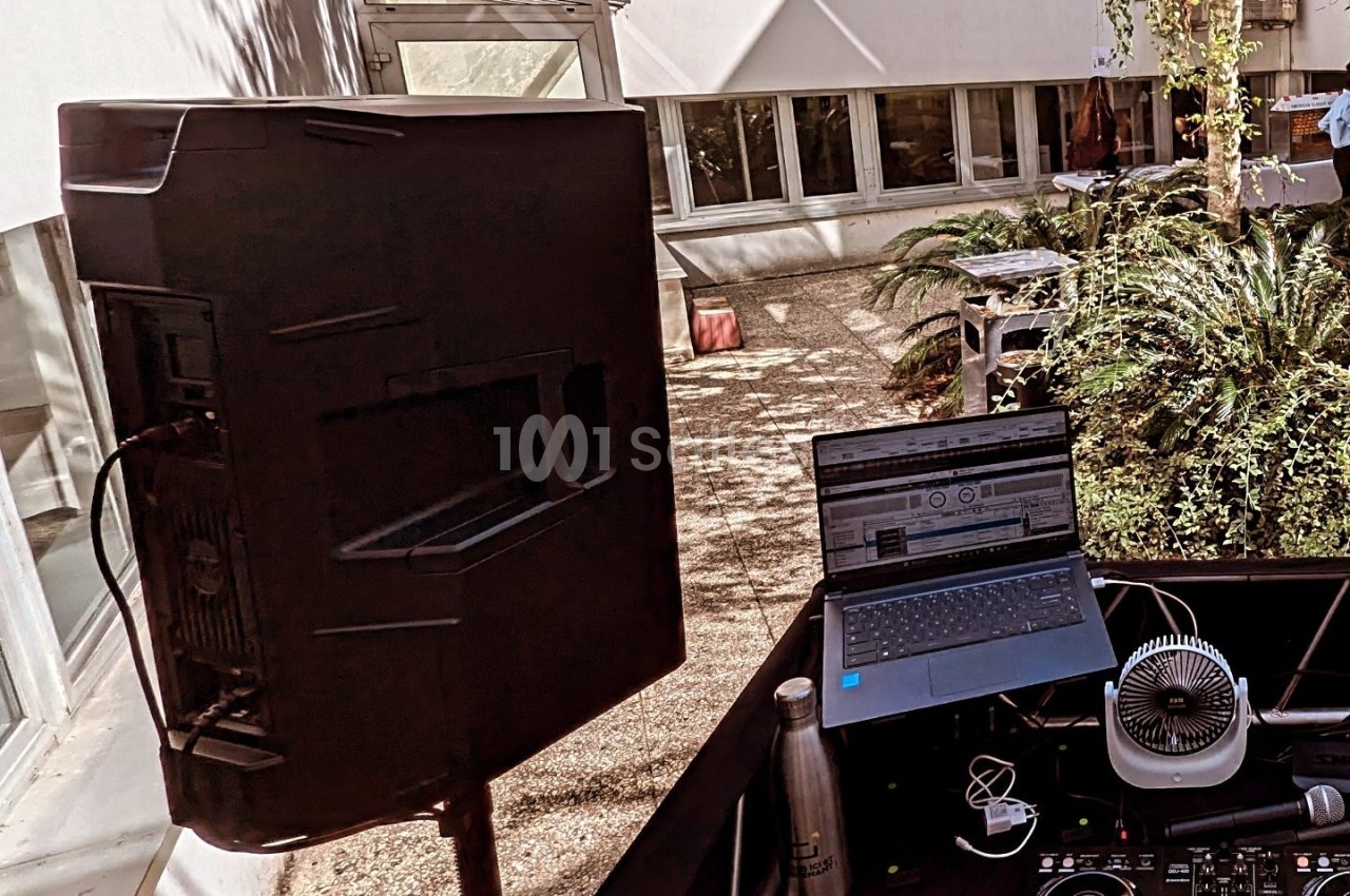 Poste de DJ en extérieur avec ordinateur portable, enceinte, ventilateur et plantes en arrière-plan.