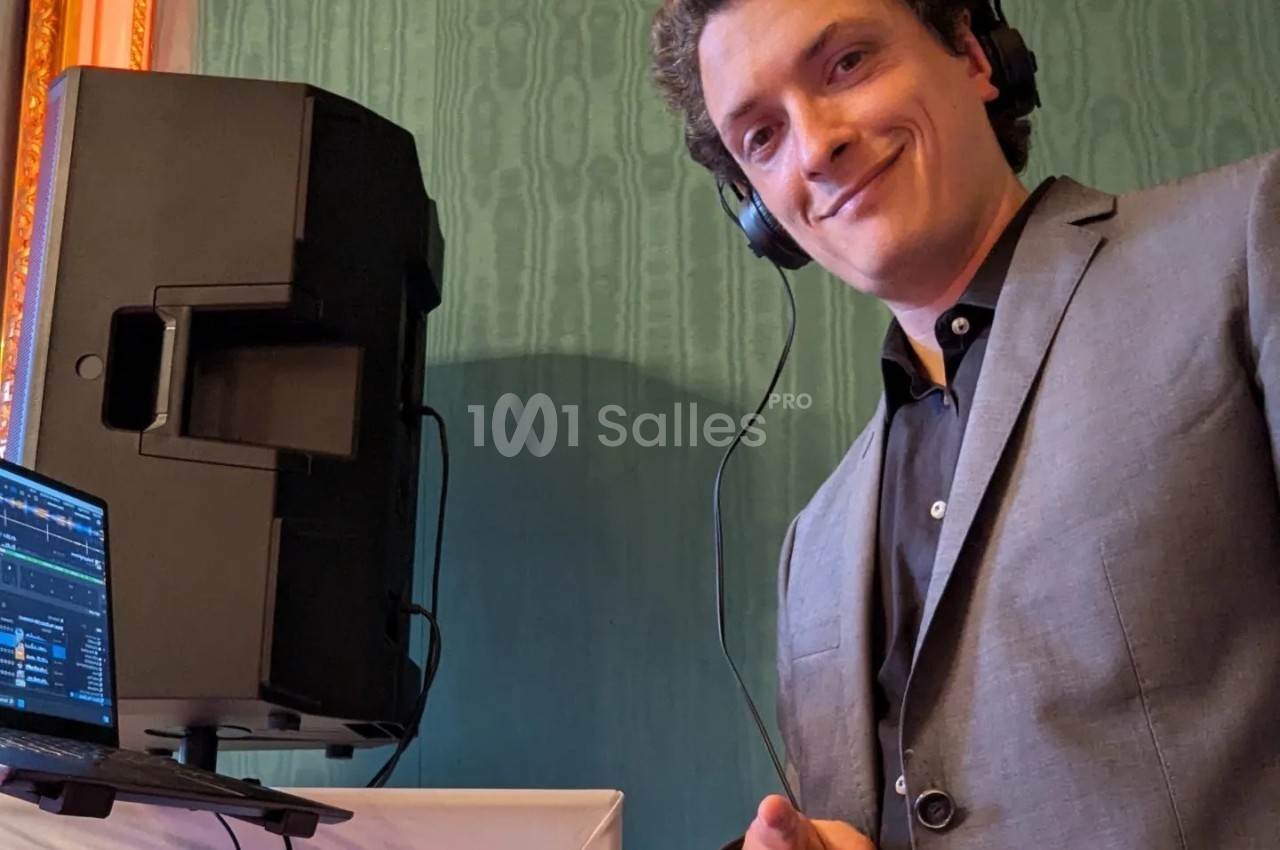 Un DJ souriant en costume mixe de la musique avec un casque, devant une enceinte et un ordinateur portable.