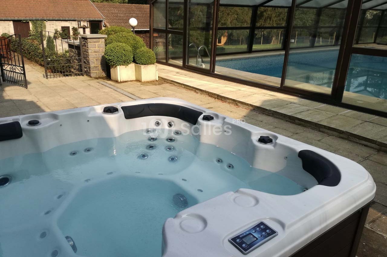 Spa extérieur avec jets massants au premier plan, près d'une piscine couverte entourée de verdure.