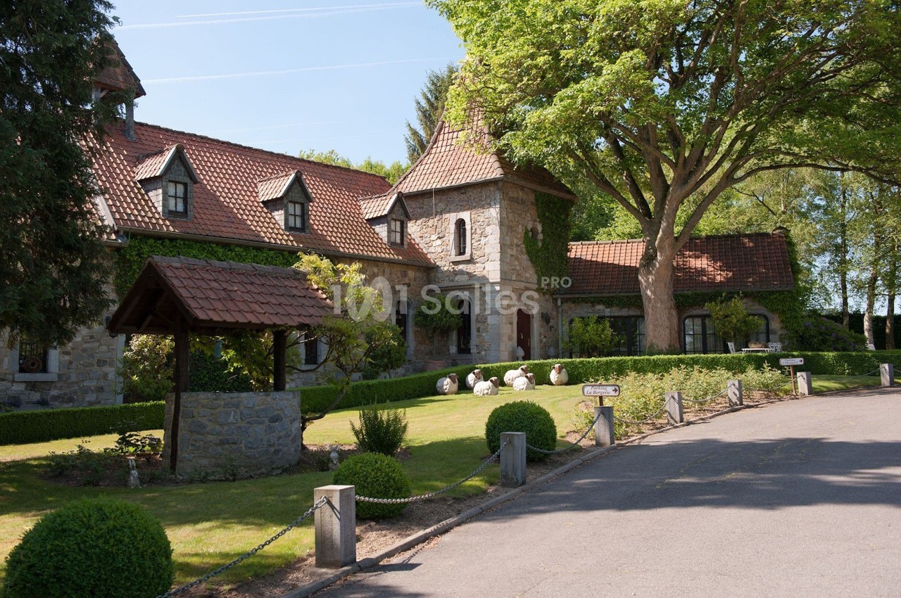Grande maison en pierre avec toit en tuiles rouges, entourée d'un jardin arboré et d'un puits en premier plan.