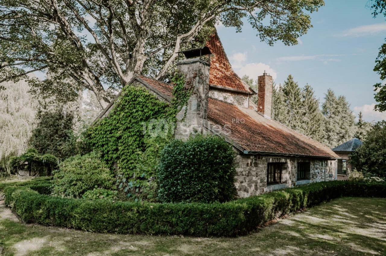 Maison en pierre entourée de verdure, avec un toit en tuiles rouges et des arbres en arrière-plan.