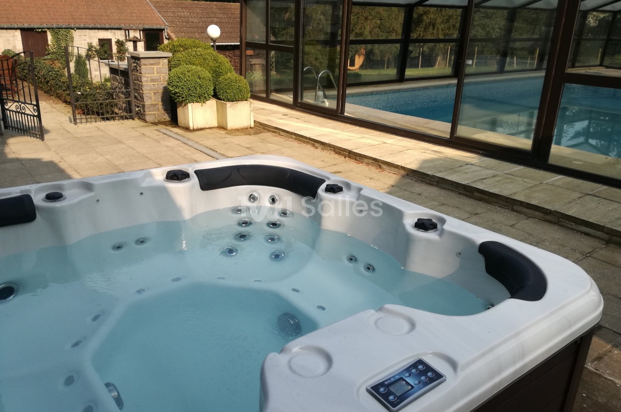 Spa extérieur avec eau claire, situé sur une terrasse près d'une piscine couverte entourée de verdure.