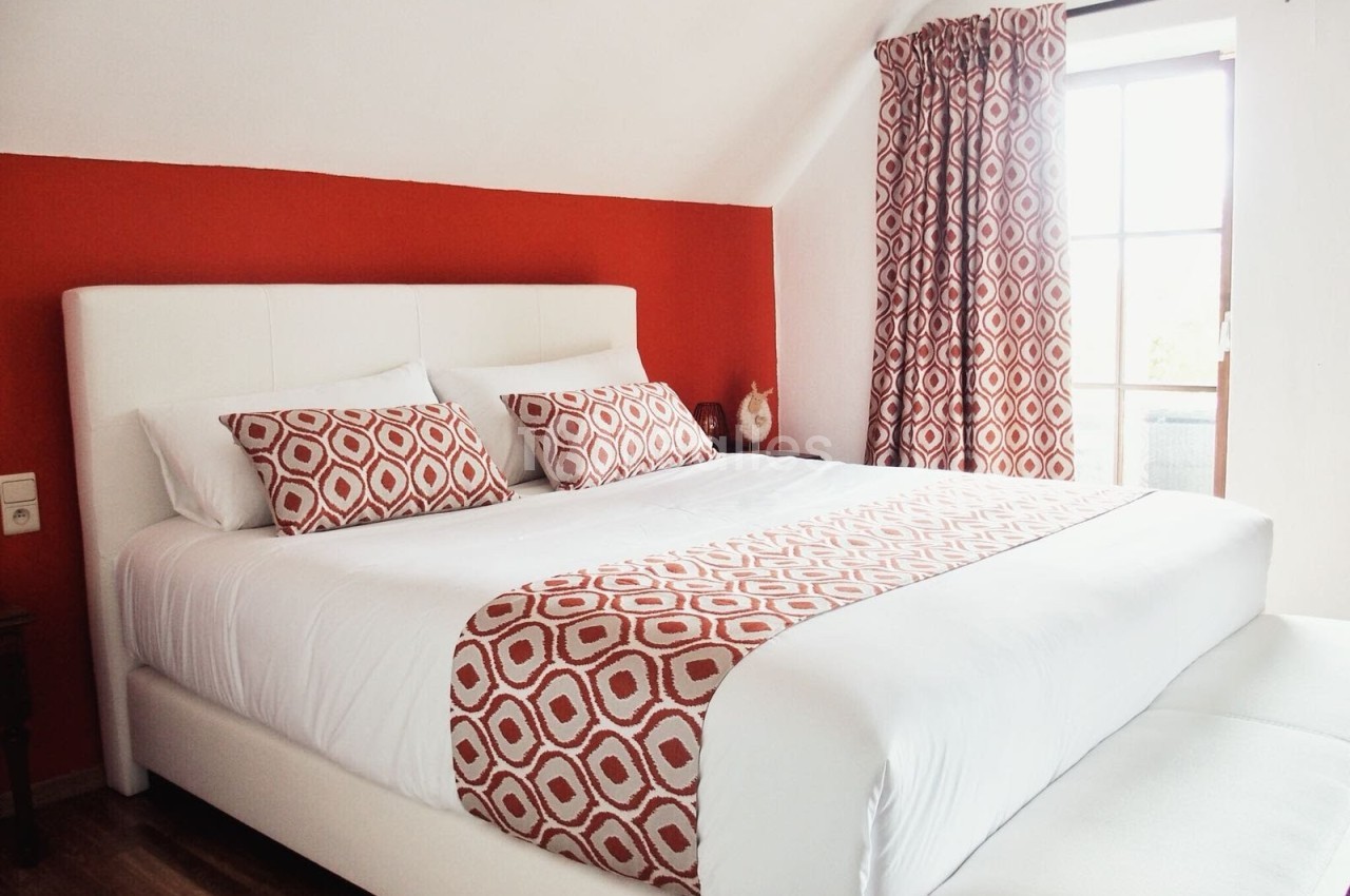 Lit double avec linge de lit blanc et motifs rouges, dans une chambre aux murs rouges et une fenêtre avec rideaux assortis.