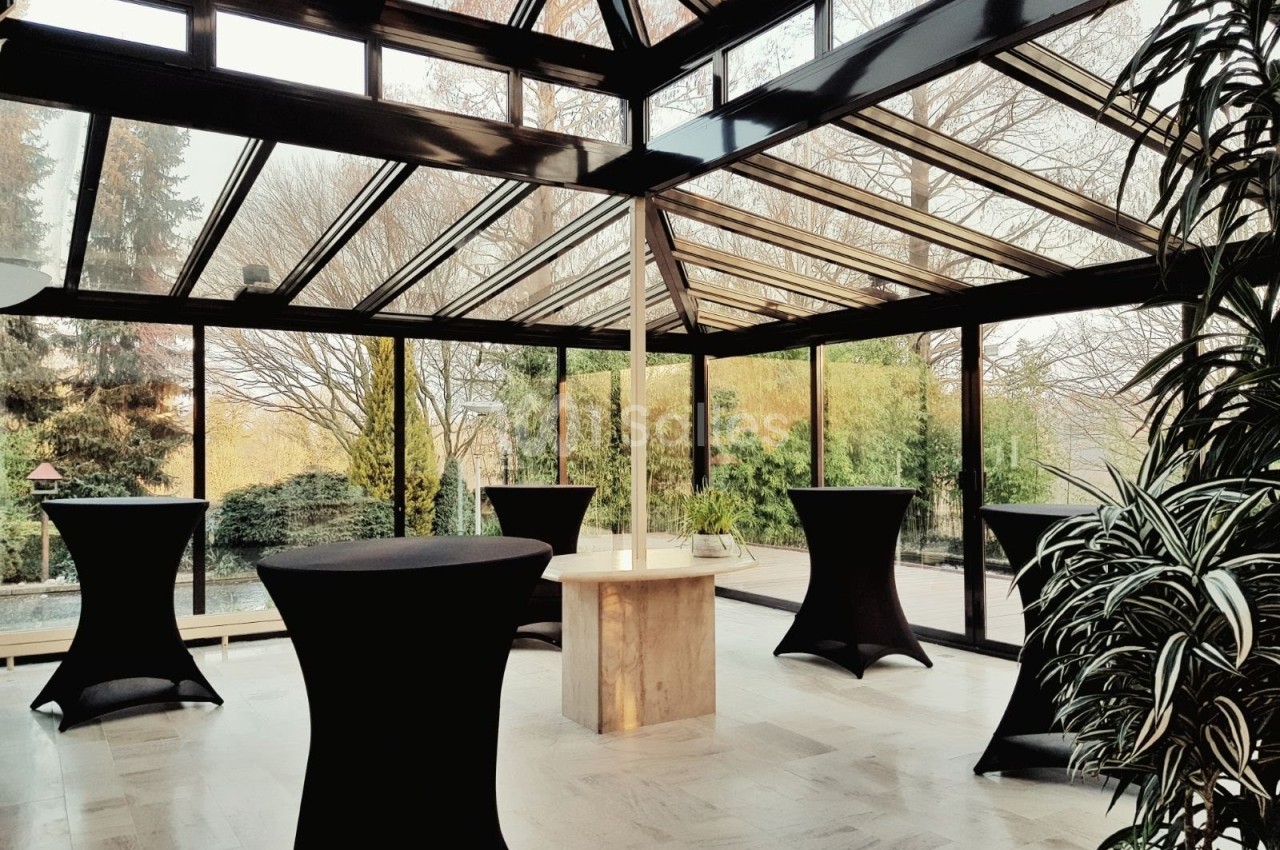 Salle lumineuse avec verrière, tables hautes noires et vue sur un jardin arboré en arrière-plan.