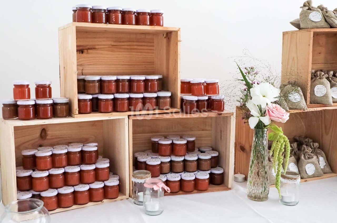 Pots de confiture disposés sur des caisses en bois, accompagnés de fleurs et de sachets en toile sur une table blanche.