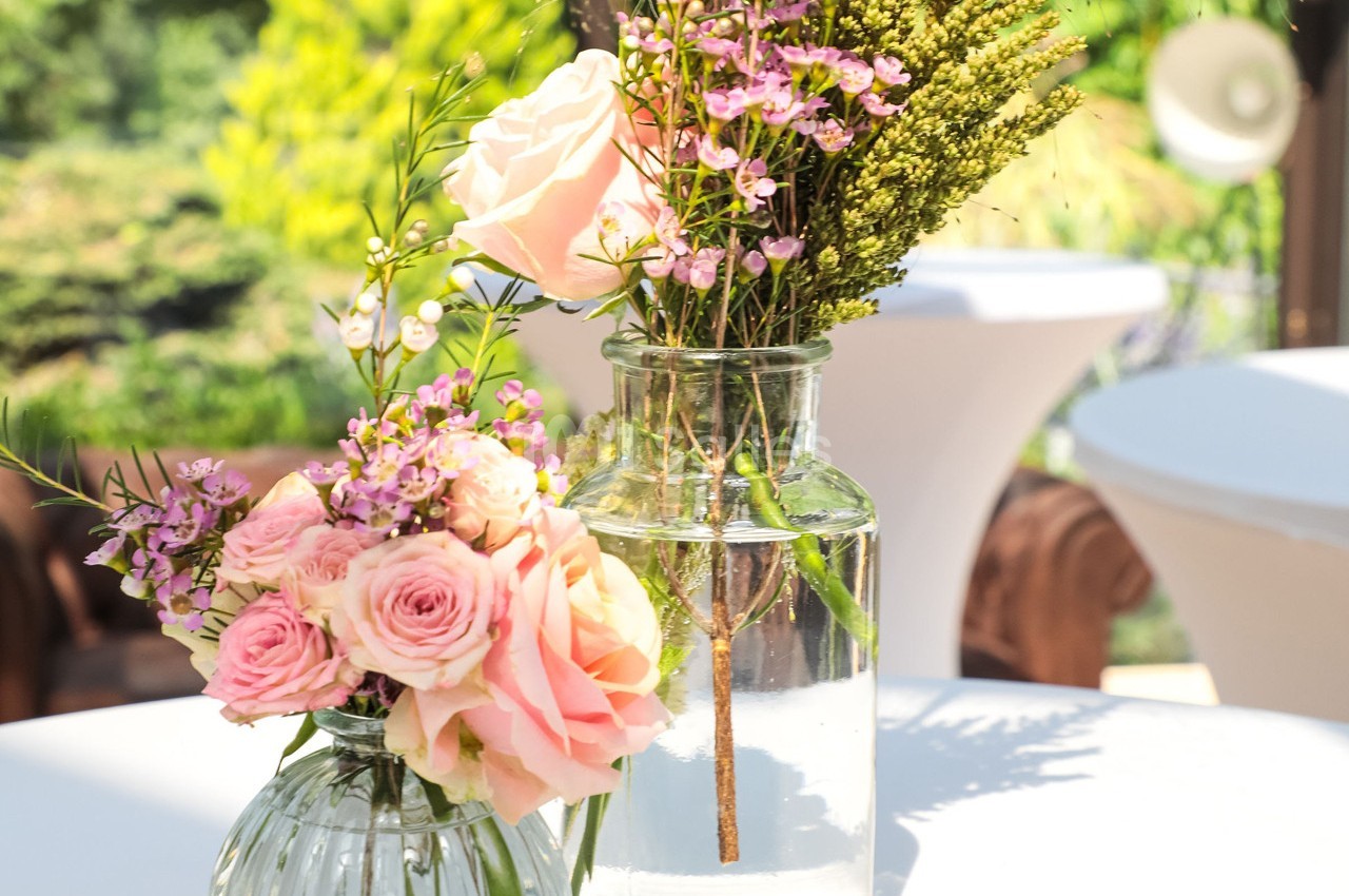 Deux vases en verre contenant des roses roses et des fleurs sauvages, posés sur une table en extérieur.