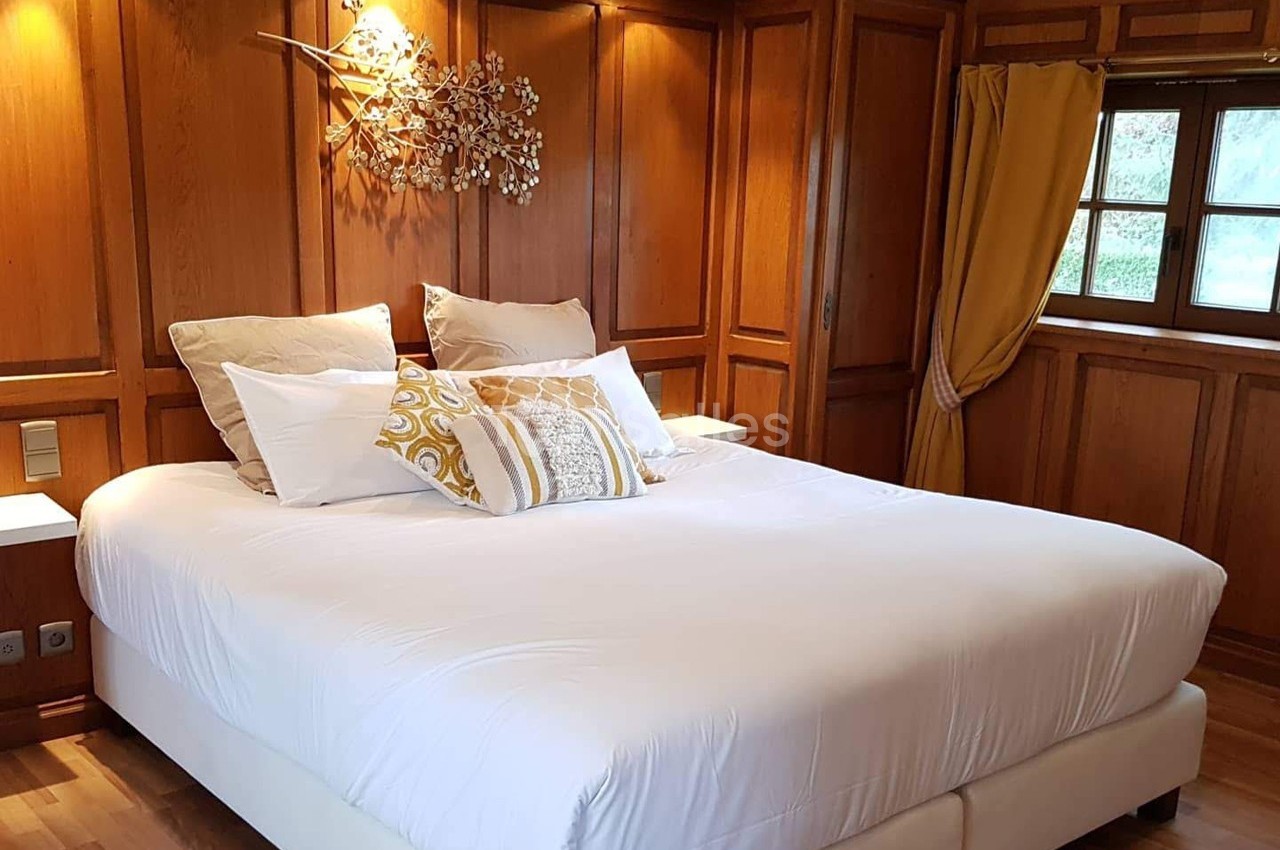 Lit double avec draps blancs, coussins décoratifs et tête de lit en bois, dans une chambre lumineuse avec fenêtre et rideaux…