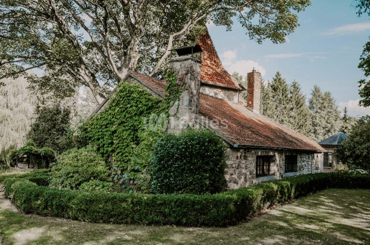 Maison en pierre entourée de verdure, avec un toit en tuiles rouges et des arbres en arrière-plan.