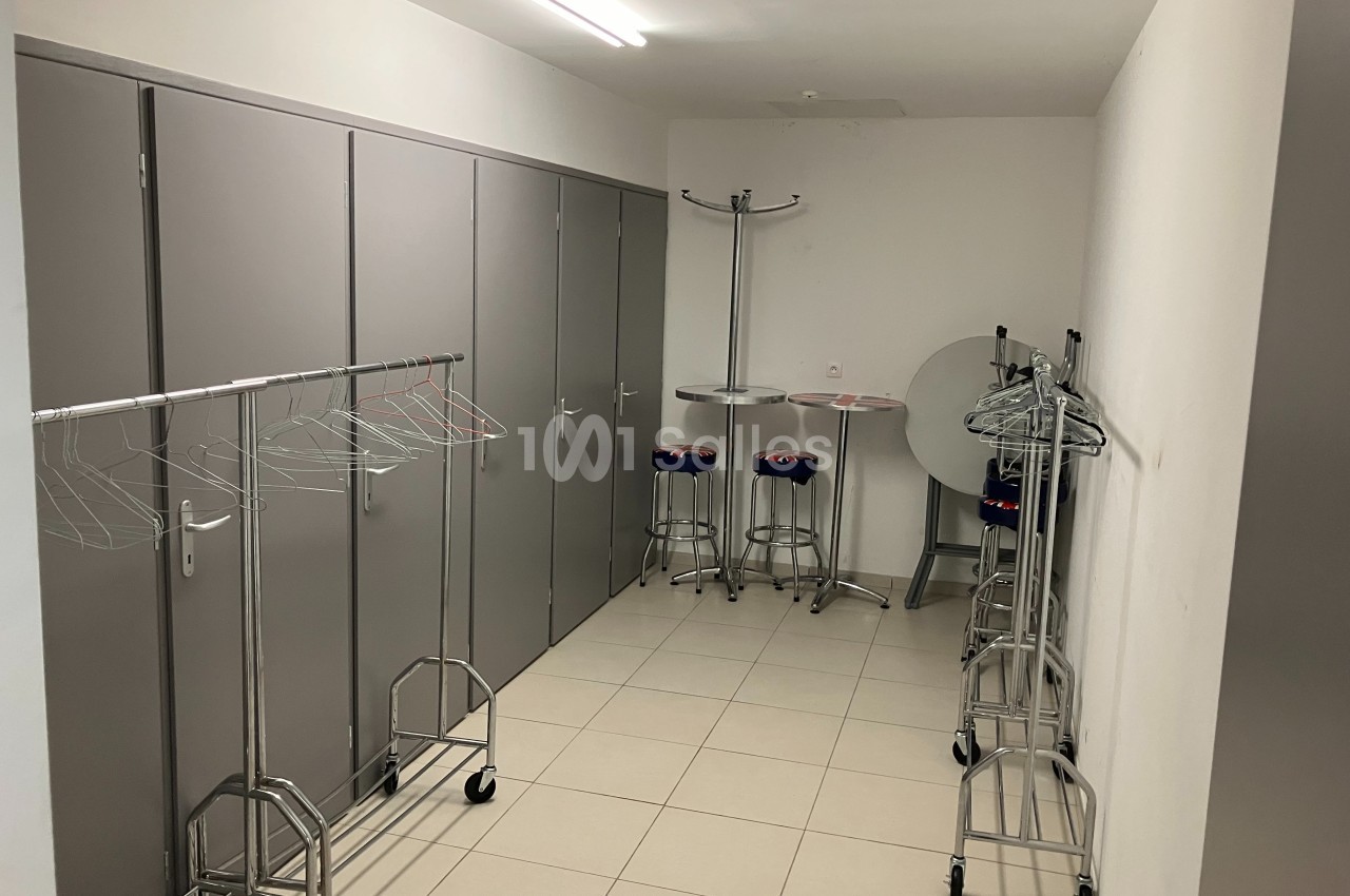 Vestiaire avec porte-manteaux sur roulettes, tabourets hauts et tables pliantes dans une pièce carrelée et éclairée.
