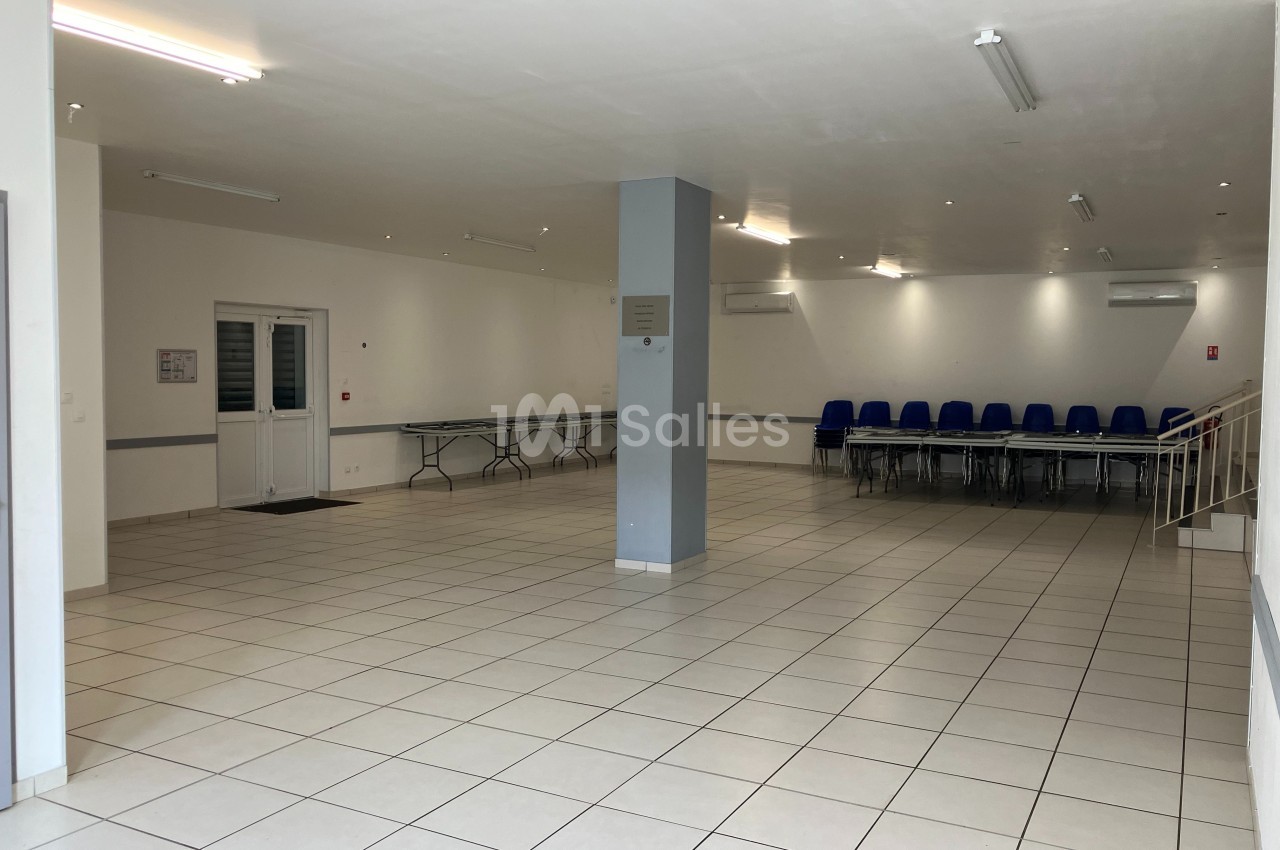 Salle vide avec carrelage blanc, chaises bleues empilées dans un coin, tables pliées le long d'un mur et éclairage au…
