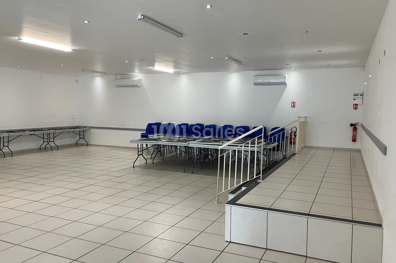Salle vide avec carrelage clair, tables pliées le long des murs, chaises empilées et une rampe d'accès.