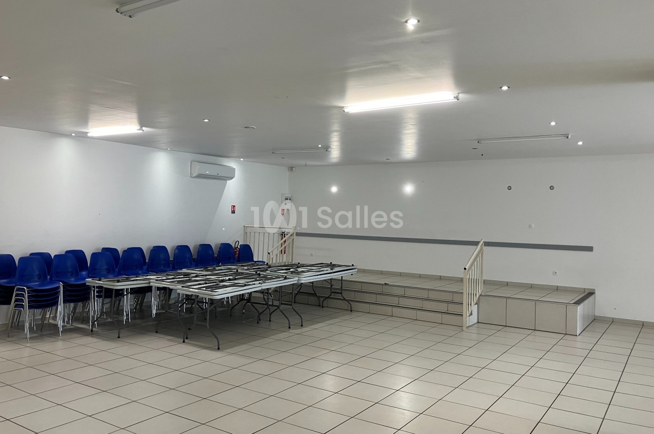 Salle vide avec sol carrelé, tables pliantes, chaises empilées à gauche et petite estrade au fond.